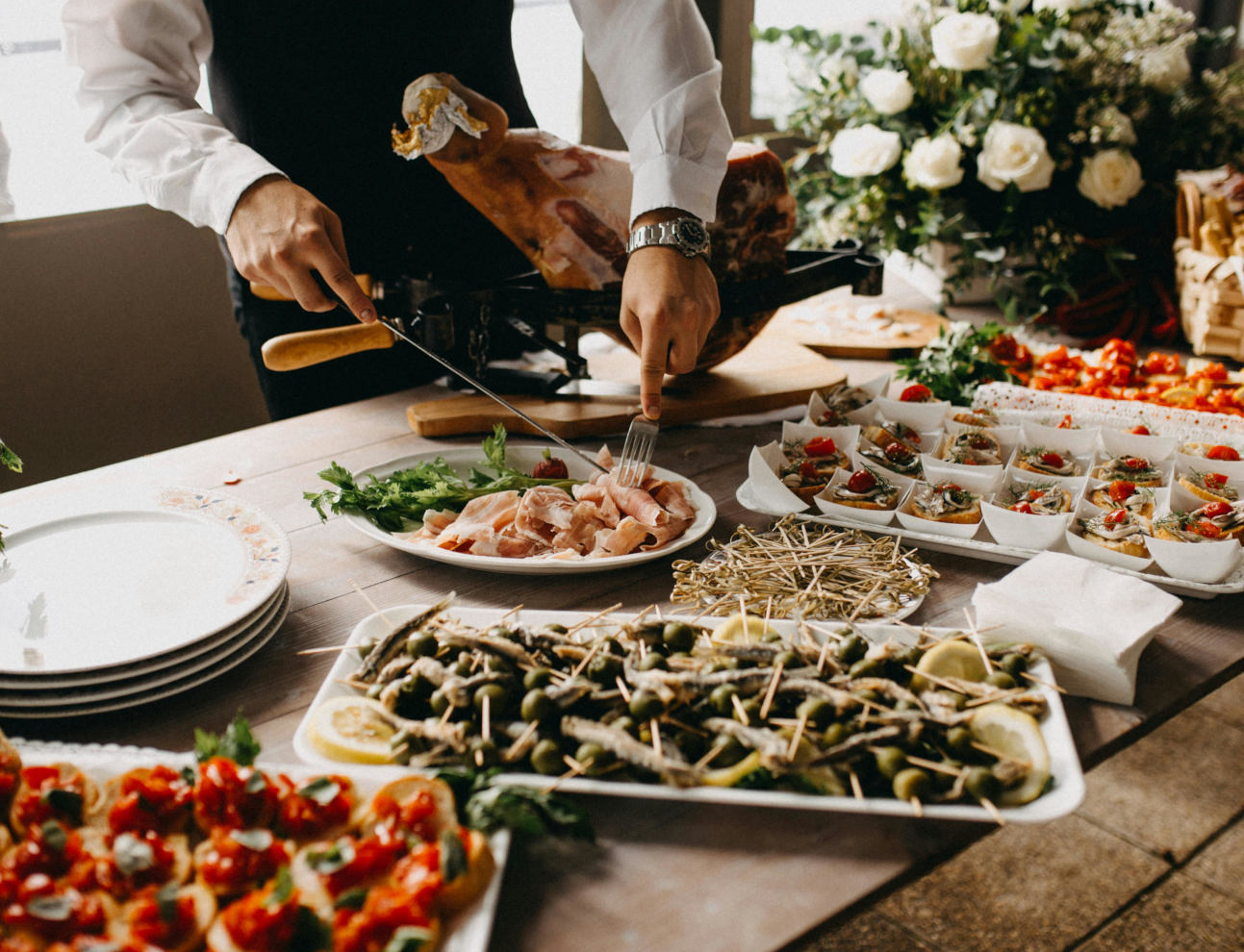 A caterer slicing cured ham beside trays of appetisers at a buffet table.